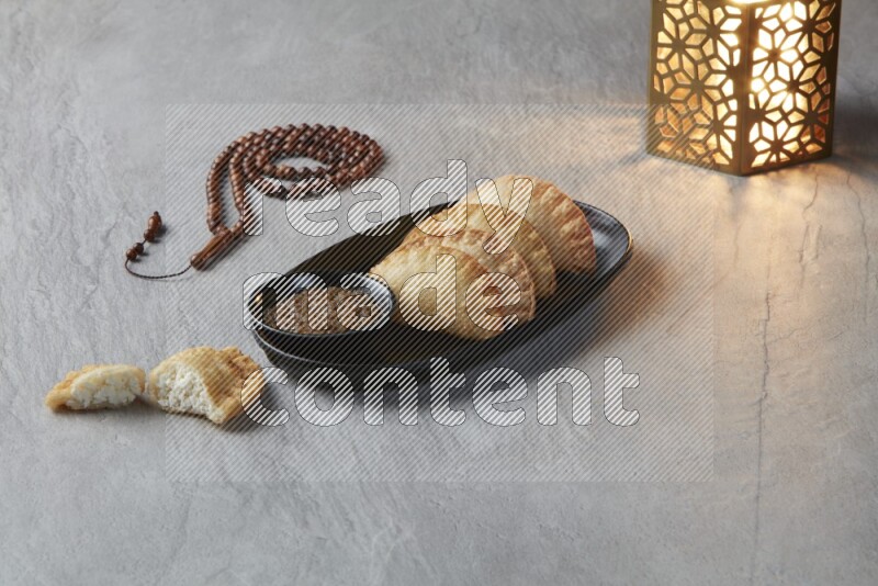 Four fried sambosas in an oval shaped black plate, beside a cut cheese sambosa, a brown misbaha and a golden lantern on a gray background