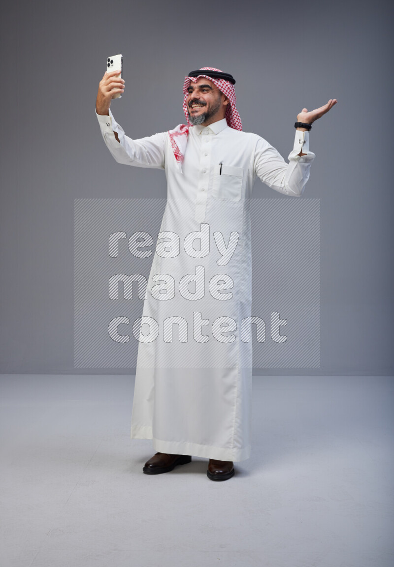 Saudi man Wearing Thob and red Shomag standing taking selfie on Gray background