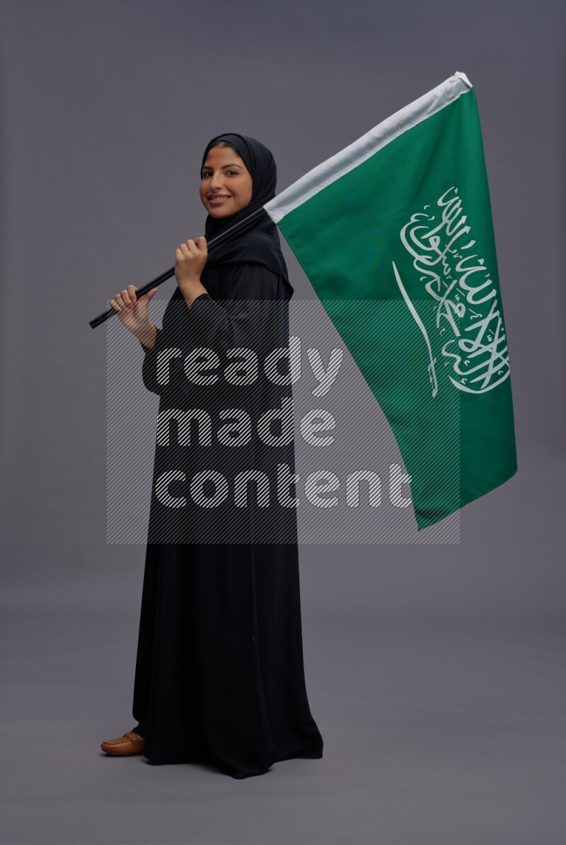 Saudi woman wearing Abaya standing holding Saudi flag on gray background