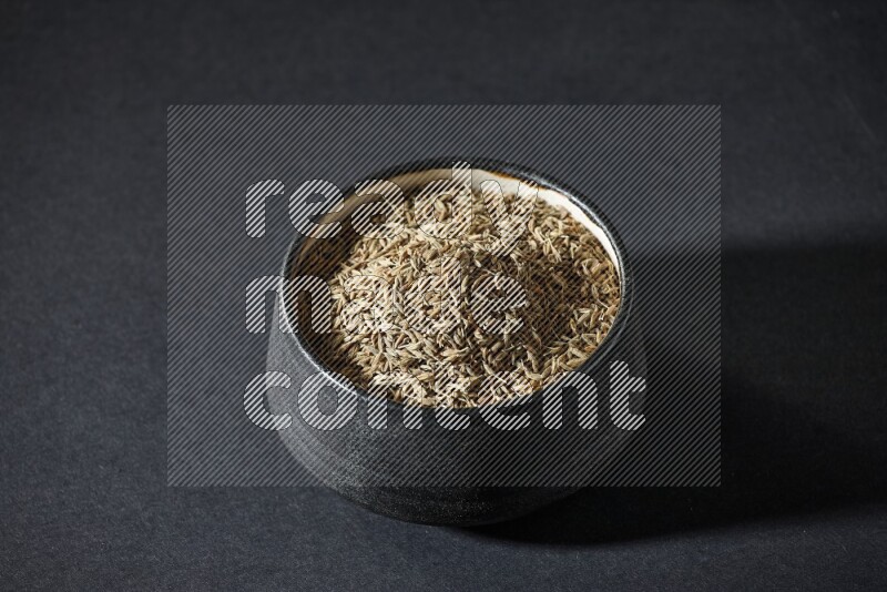A black pottery bowl full of cumin seeds on a black flooring