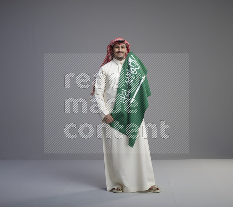 A saudi man standing wearing thob and red shomag holding big saudi flag on gray background