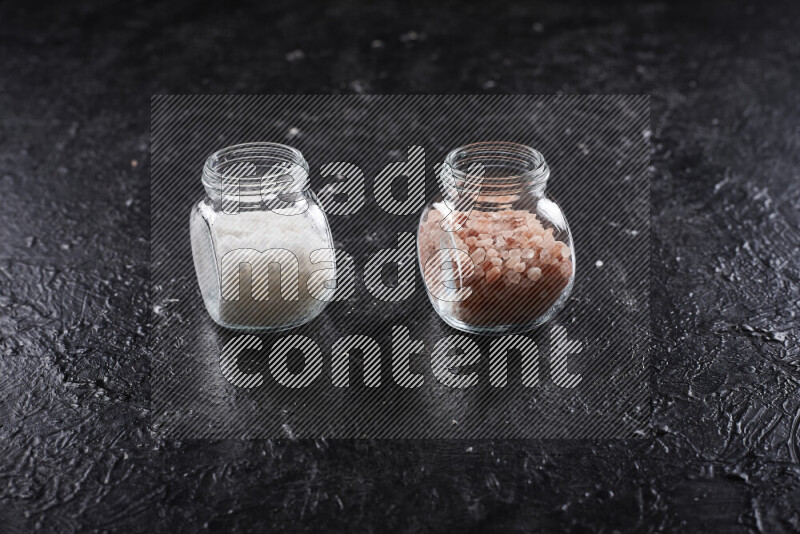 2 glass jars one is filled with coarse himalayan salt and the other with coarse sea salt on black background