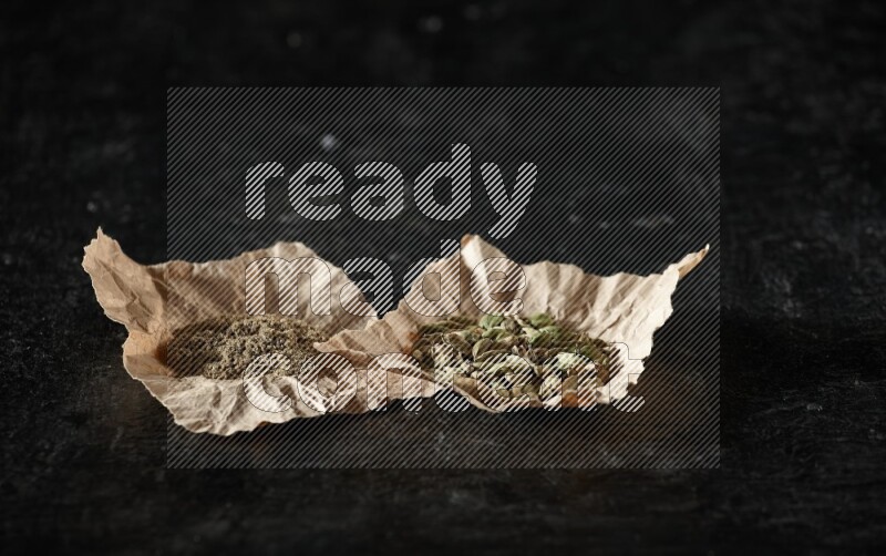 Cardamom seeds and cardamom powder in 2 crumpled pieces of paper on textured black flooring