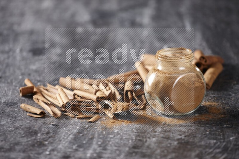 Herbal glass jar and a metal spoon full of cinnamon powder surrounded by cinnamon sticks on textured black background