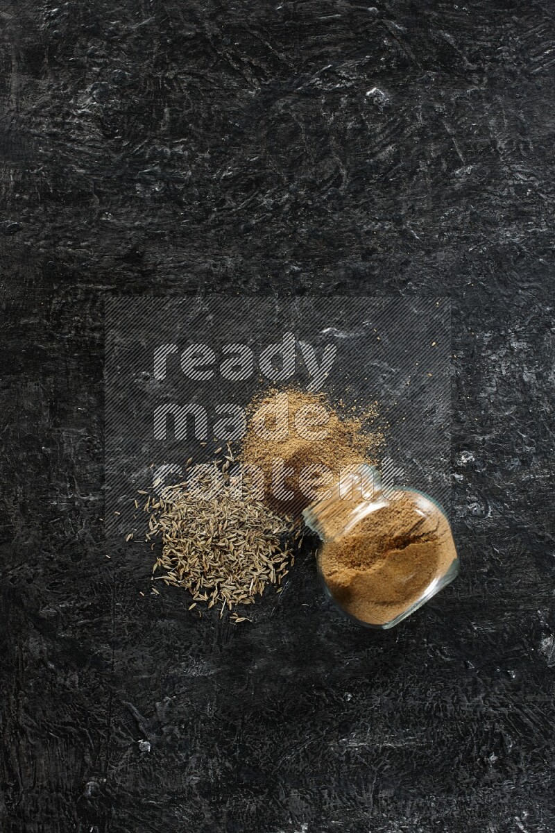 A flipped glass spice jar full of cumin powder with spilled powder and cumin seeds on a textured black flooring