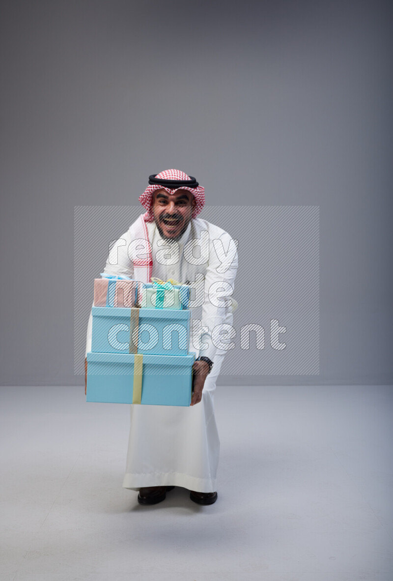 Saudi man Wearing Thob and red Shomag standing holding gift box on Gray background