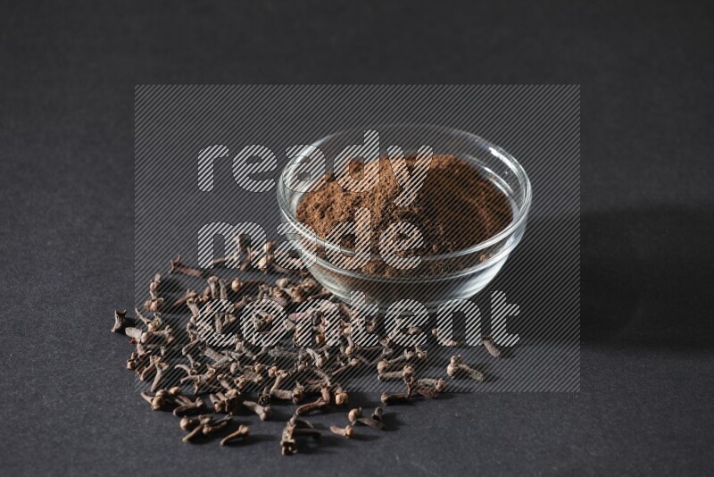 A glass bowl full of cloves powder with cloves grains spread on black flooring