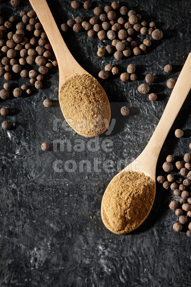 Wooden spoons full of all spice powder and allspice whole balls beside it on a textured black flooring