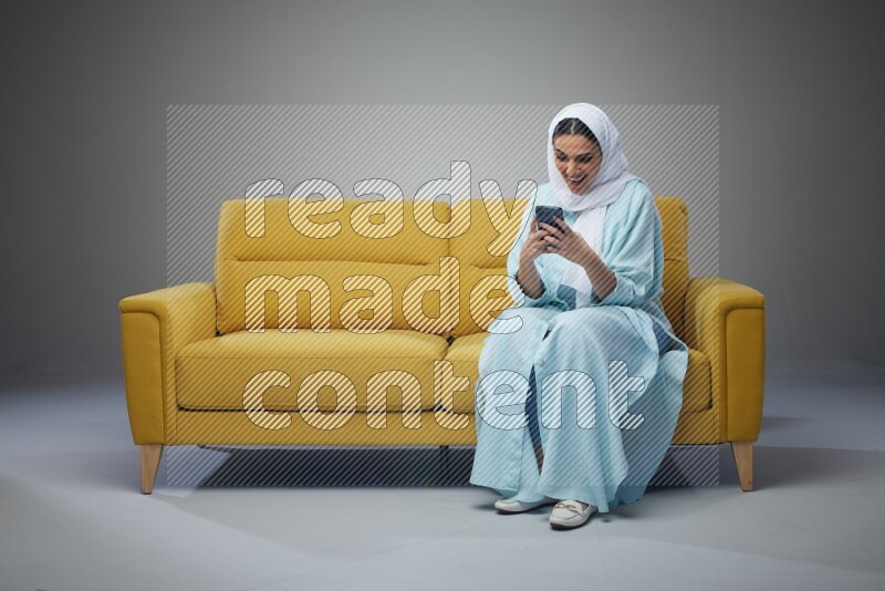 A Saudi woman wearing a light blue Abaya and white head scarf sitting on a yellow sofa and texting on her phone on a grey background
