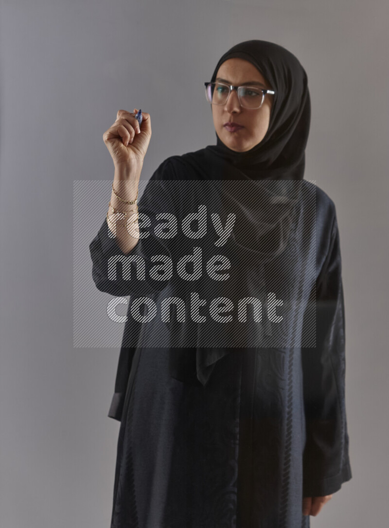 Woman in black abaya, hijab and glasses holding a marker pen to write on a transparent board with different reactions and expressions on grey background