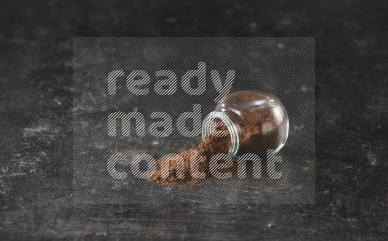 A flipped glass spice jar full of cloves powder and powder came out of it on textured black flooring