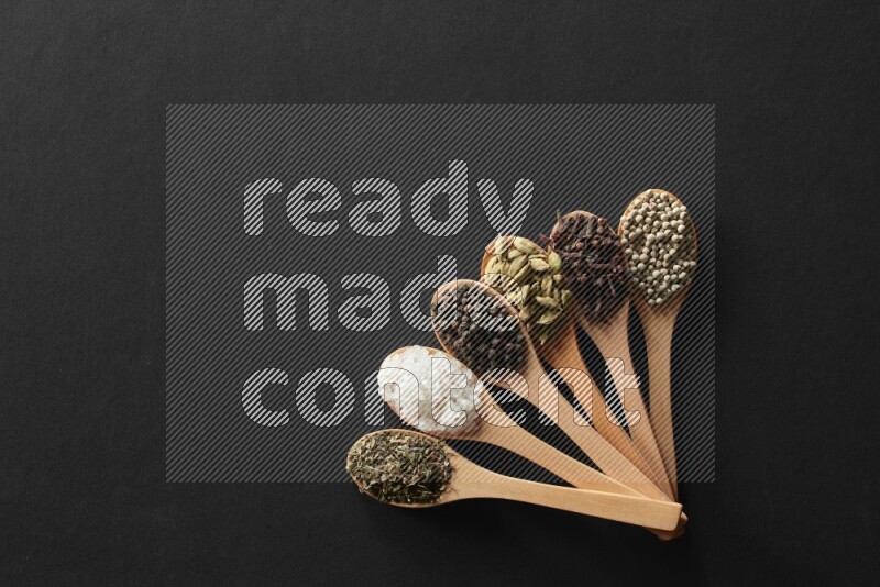 الفلفل الأبيض والقرنفل والهيل والملح والفلفل الأسود وريحان مجفف في معالق خشبية علي خلفية سوداء