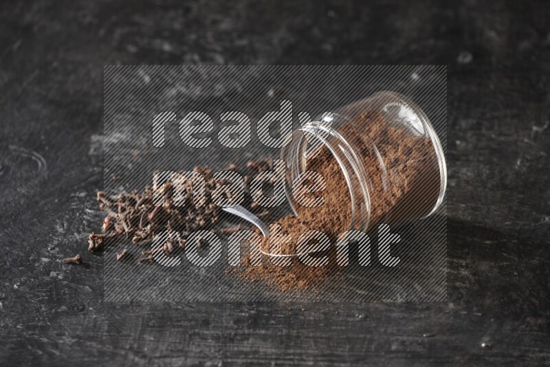 A flipped glass jar and metal spoon full of cloves powder with cloves spread on a textured black flooring