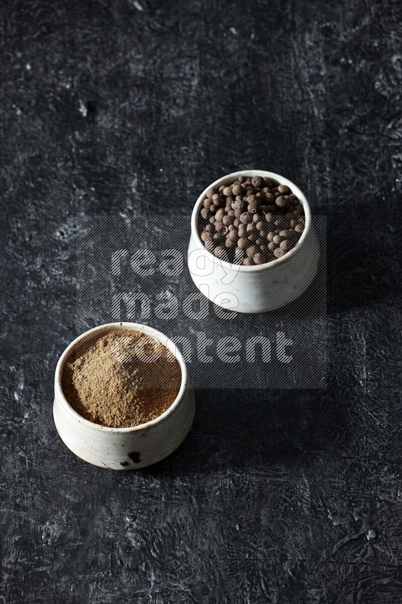2 beige bowls, one full of allspice powder and the other full of whole balls on a textured black flooring