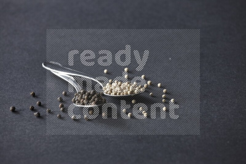 ملعقتان معدنيتان ممتلئتان بحبوب الفلفل الأسود والأبيض مع حبوب متناثرة على أرضية سوداء