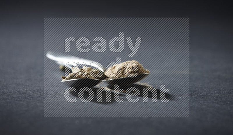 2 Metal spoons full of cardamom powder and cardamom seeds on black flooring