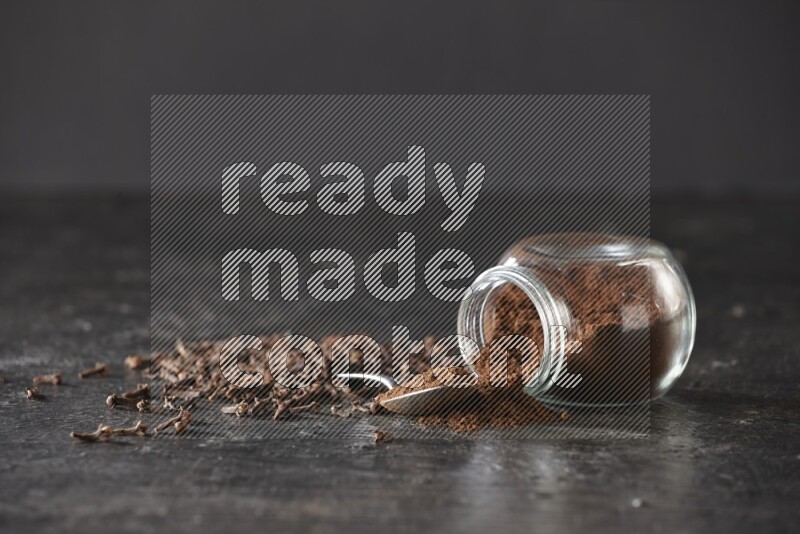 A flipped glass spice jar and a metal spoon full of cloves powder with cloves spread on textured black flooring