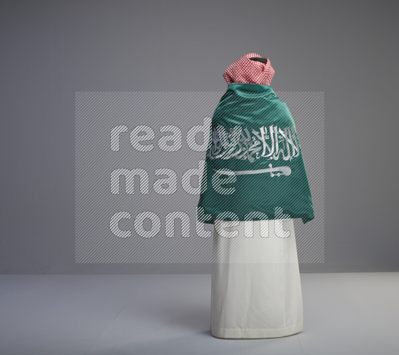 A Saudi man standing wearing thob and red shomag wrapping big saudi flag on gray background