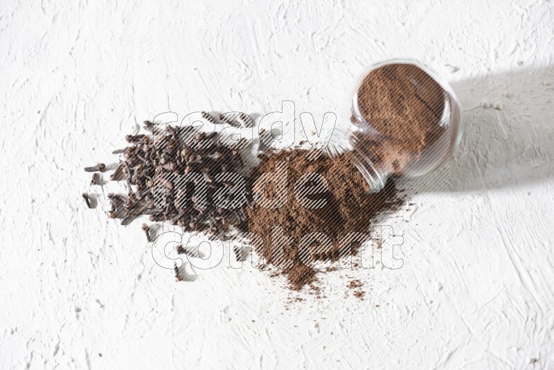 وعاء زجاجي للتوابل مقلوب ممتلئ ببودرة القرنفل وتناثرت البودرة منه مع حبوب القرنفل المنتشرة على أرضية بيضاء