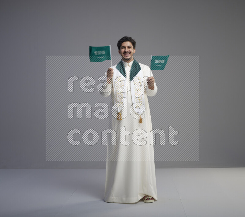 A Saudi man standing wearing thob and saudi flag scarf and holding small saudi flag on gray background