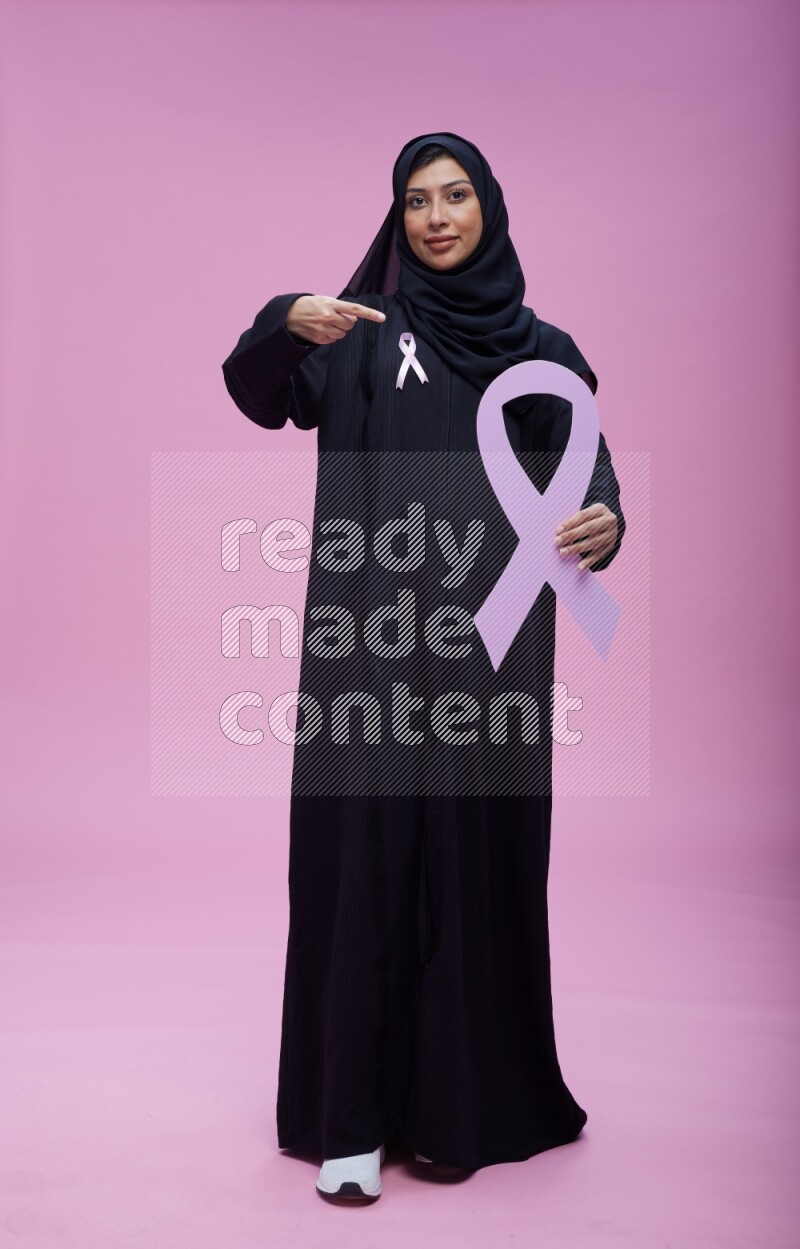 Saudi woman wearing pink ribbon on abaya standing holding awareness ribbon on plain pink background