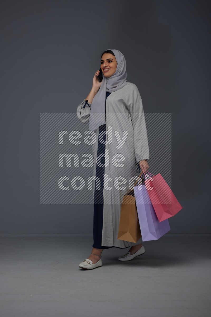 A Saudi woman wearing a light gray Abaya and head scarf standing and holding shopping bags making different poses eye level on a grey background