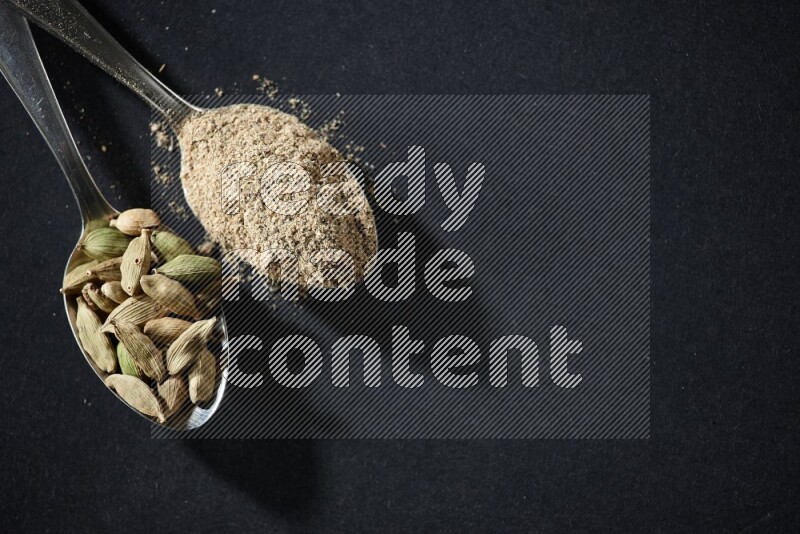 ملعقتان معدنيتان ممتلئتان ببودرة الهيل وحبوب الهيل على خلفية سوداء