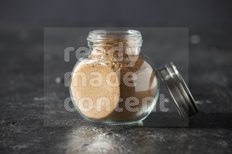 A glass spice jar full of allspice powder on a textured black flooring