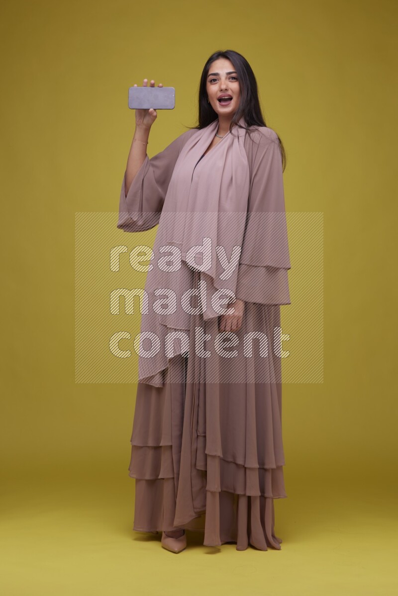 A woman Showing SmartPhone Screen on a Yellow Background wearing Brown Abaya
