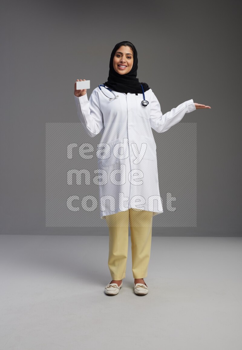 Saudi woman wearing lab coat with stethoscope standing holding ATM card on Gray background