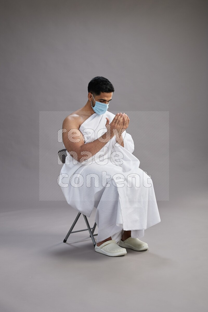 A man wearing Ehram with face mask sitting on chair dua'a on gray background