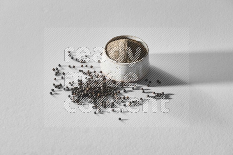 A white ceramic bowl full of black pepper powder and black pepper beads spread on white flooring