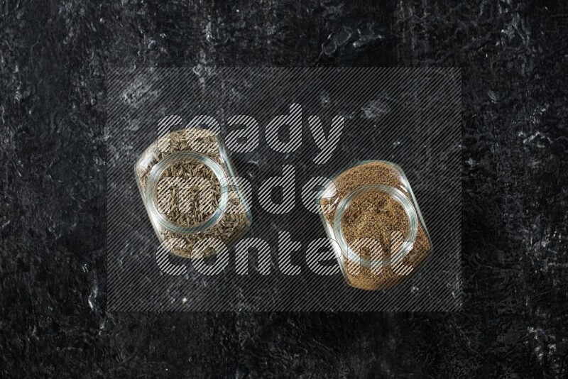 2 glass spice jars full of cumin powder and seeds on a textured black flooring