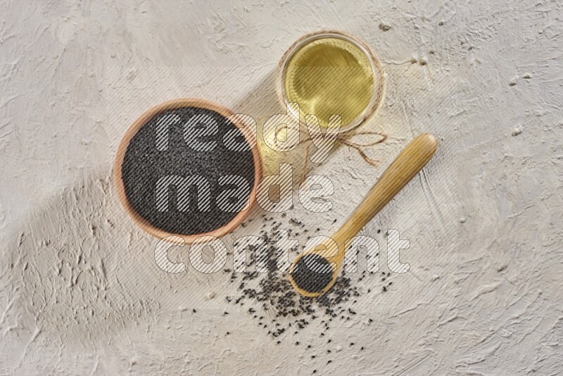 A wooden bowl and spoon full of black seeds with a glass jar of black seeds oil on a textured white flooring