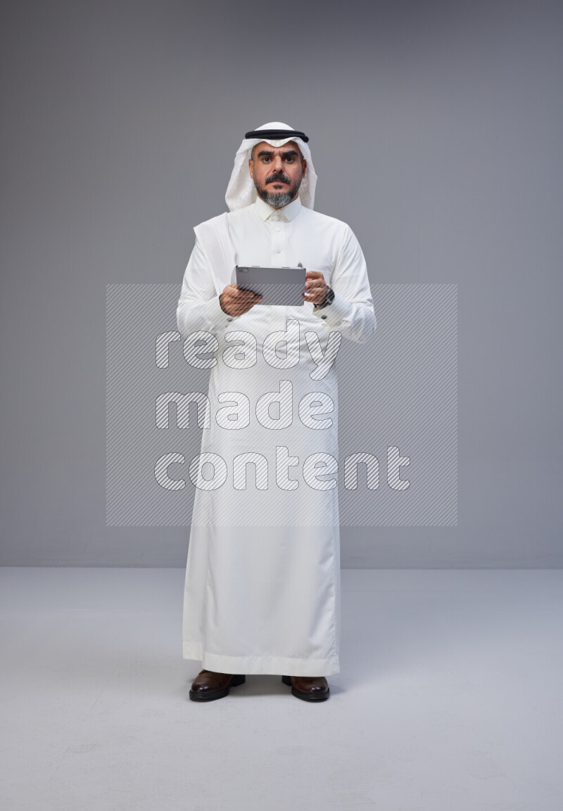 Saudi man Wearing Thob and white Shomag standing working on tablet on Gray background