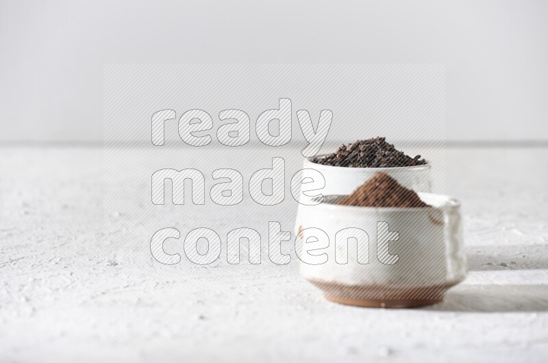 2 Beige ceramic bowls, one full of cloves and the other full of powder on textured white flooring