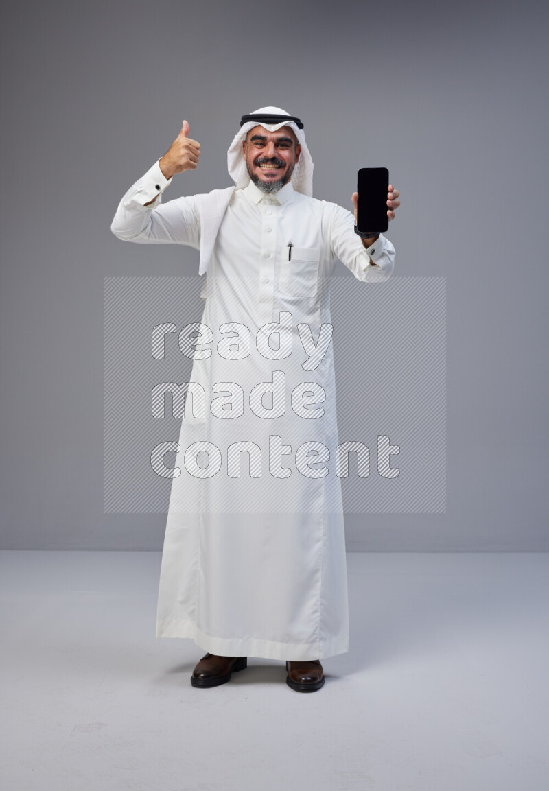 Saudi man Wearing Thob and white Shomag standing showing phone to camera on Gray background