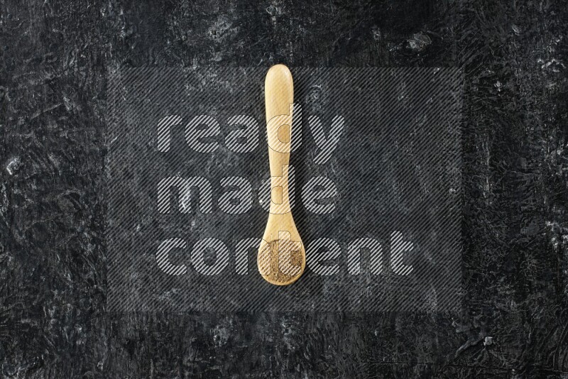 A wooden spoons full of cumin powder on textured black flooring