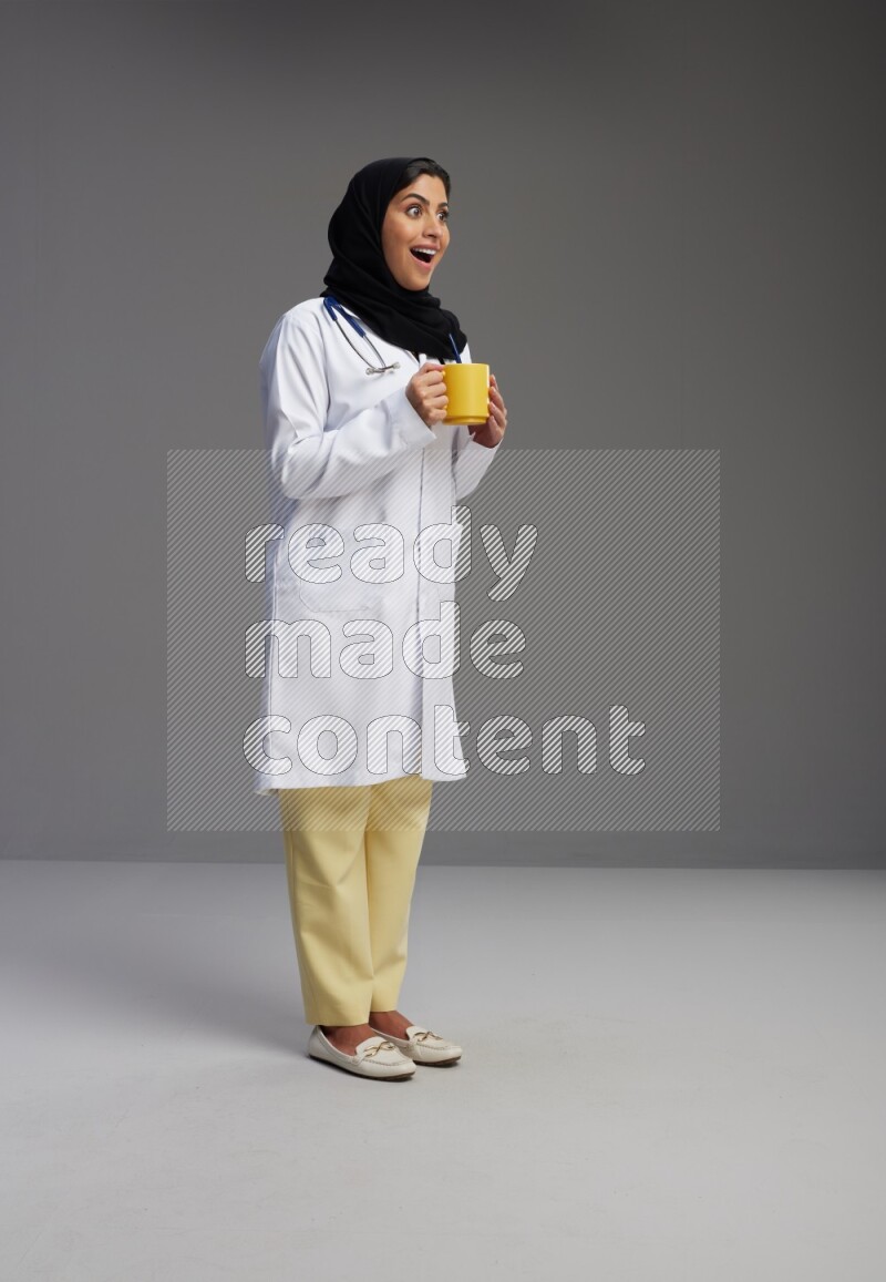 Saudi woman wearing lab coat with stethoscope standing holding mug on Gray background