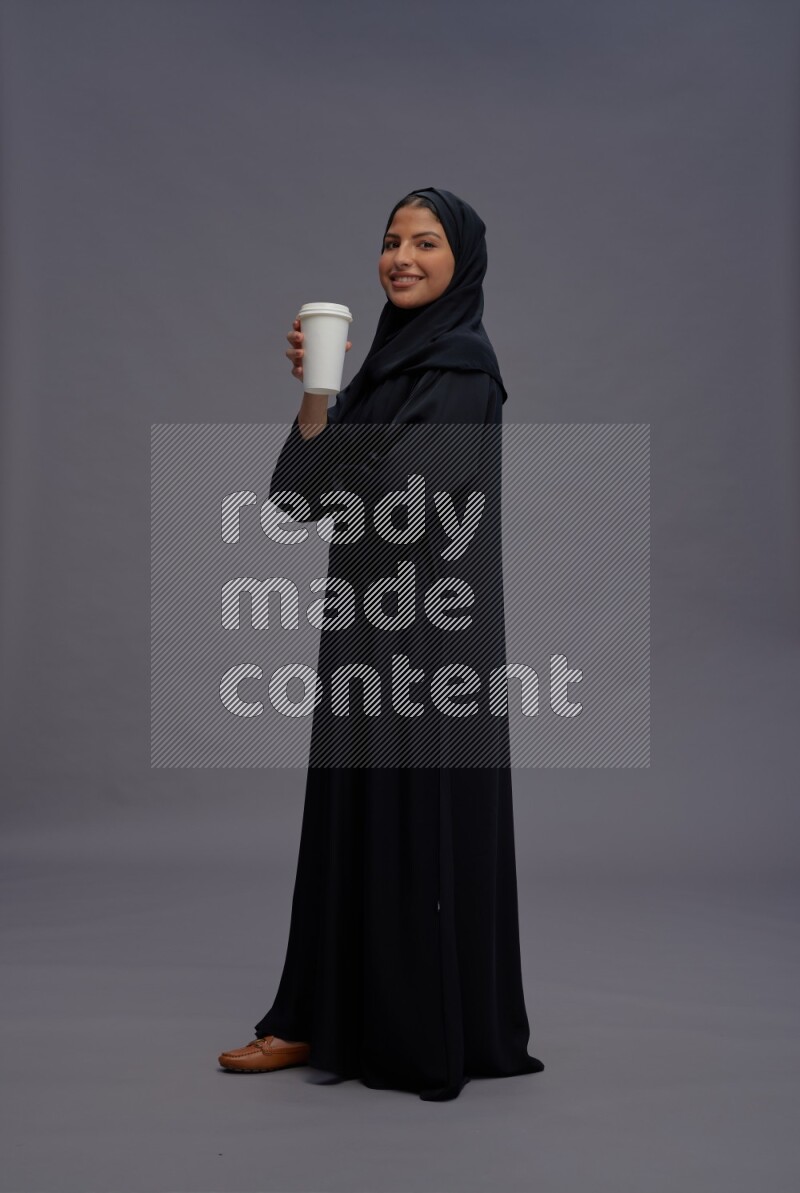 Saudi woman wearing Abaya standing holding paper cup on gray background