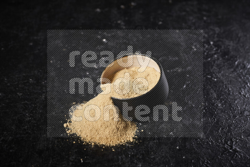 A black pottery bowl full of ground ginger powder with fallen powder from it on black background