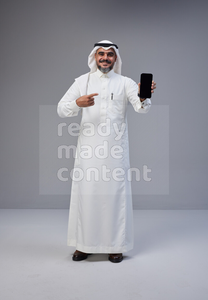 Saudi man Wearing Thob and white Shomag standing showing phone to camera on Gray background
