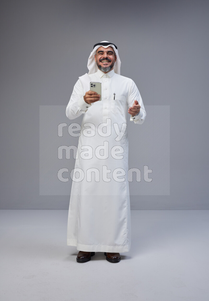 Saudi man Wearing Thob and white Shomag standing texting on phone on Gray background