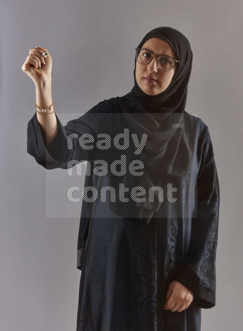 Woman in black abaya, hijab and glasses holding a marker pen to write on a transparent board with different reactions and expressions on grey background