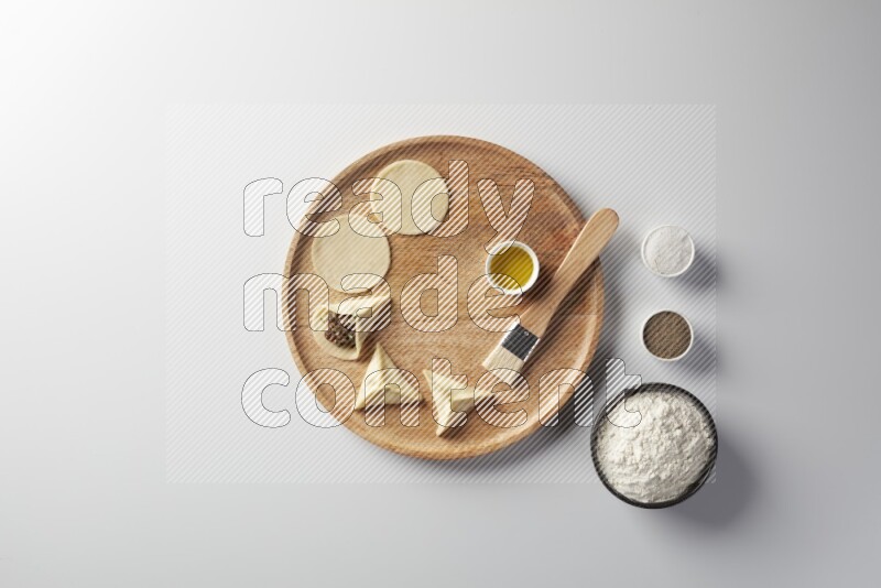 two closed sambosas and one open sambosa filled with meat while flour, salt, black pepper and oil with oil brush aside in a wooden dish on a white background