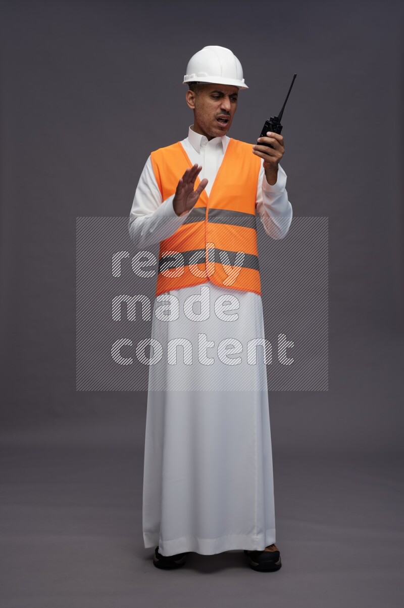 Saudi man wearing thob with engineer vest standing holding walkie-talkie on gray background