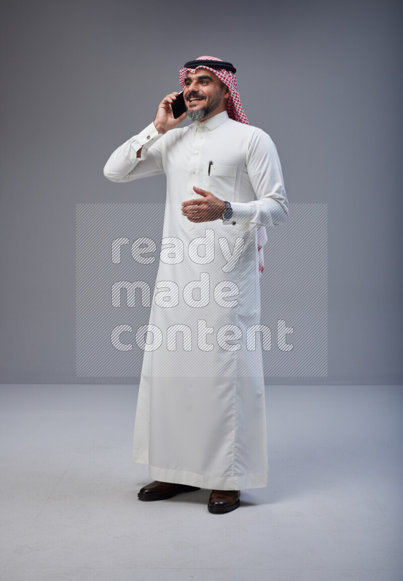 Saudi man Wearing Thob and red Shomag standing talking on phone on Gray background