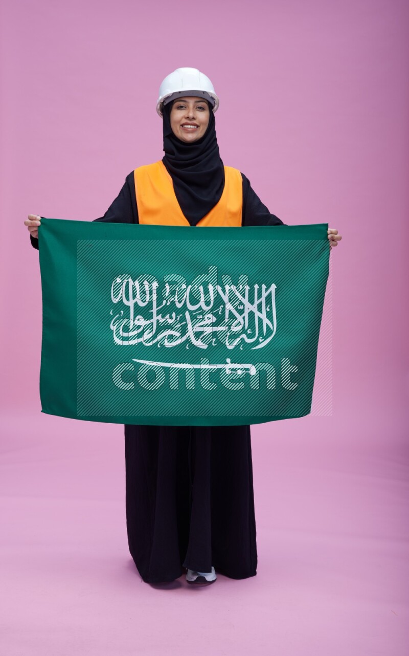 Saudi woman wearing Abaya with engineer vest and helmet standing holding Saudi flag on pink background