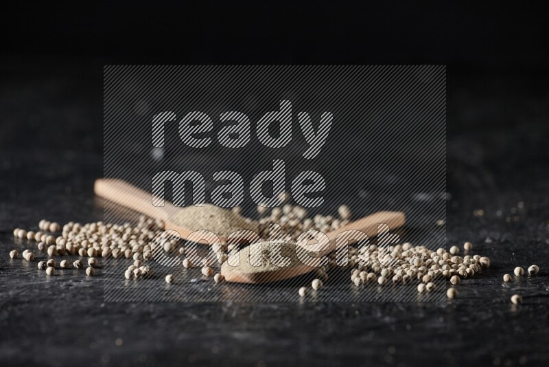 2 wooden spoons full of white pepper powder with white pepper beads on textured black flooring