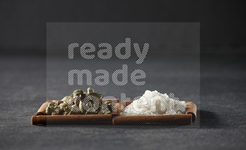 مربعان من أعواد القرفة ممتلئان بالملح الأبيض وحبوب الهيل علي خلفية سوداء